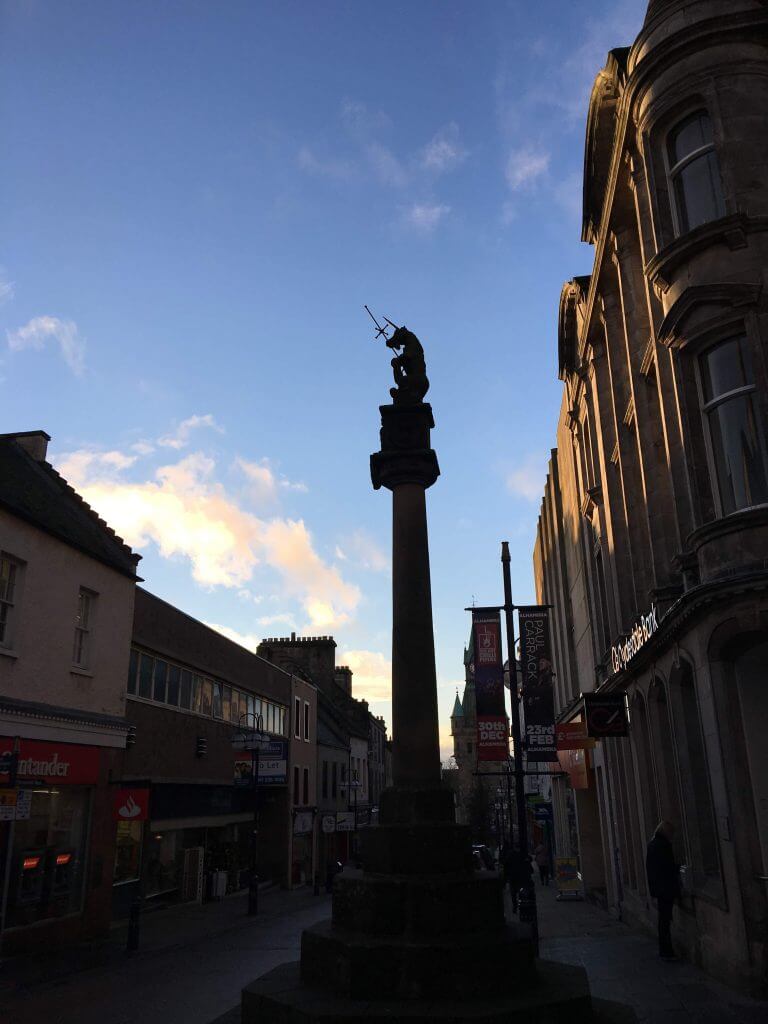 Photo of Dunfermline High Street looking west with Mercat cross, topped with Unicorn, surrounded by high street businesses. 